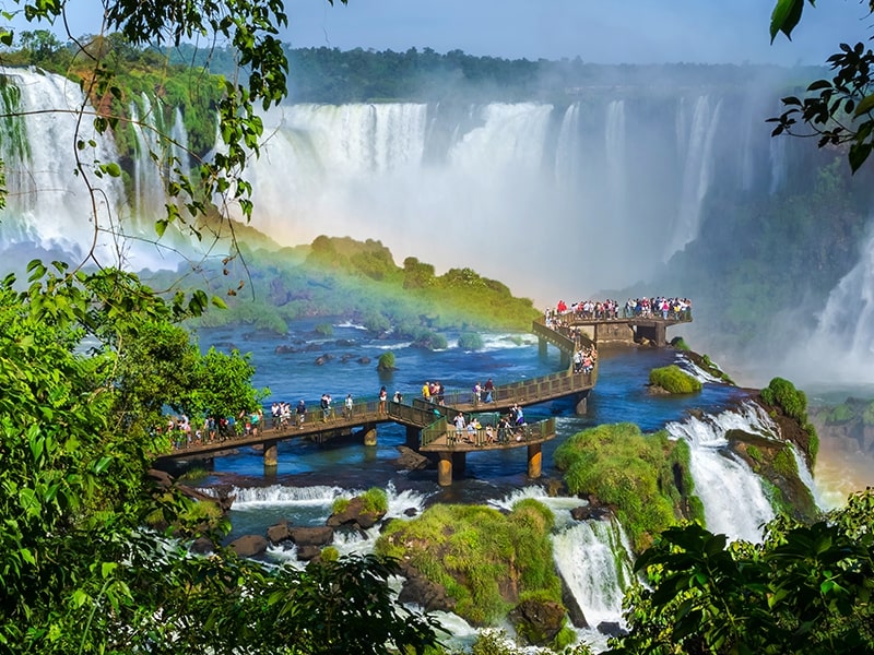 Plataforma de observação nas Cataratas do Iguaçu, onde turistas apreciam a vista deslumbrante das quedas d'água e um arco-íris que se forma com a névoa, proporcionando uma experiência única em meio à natureza de Foz do Iguaçu.
