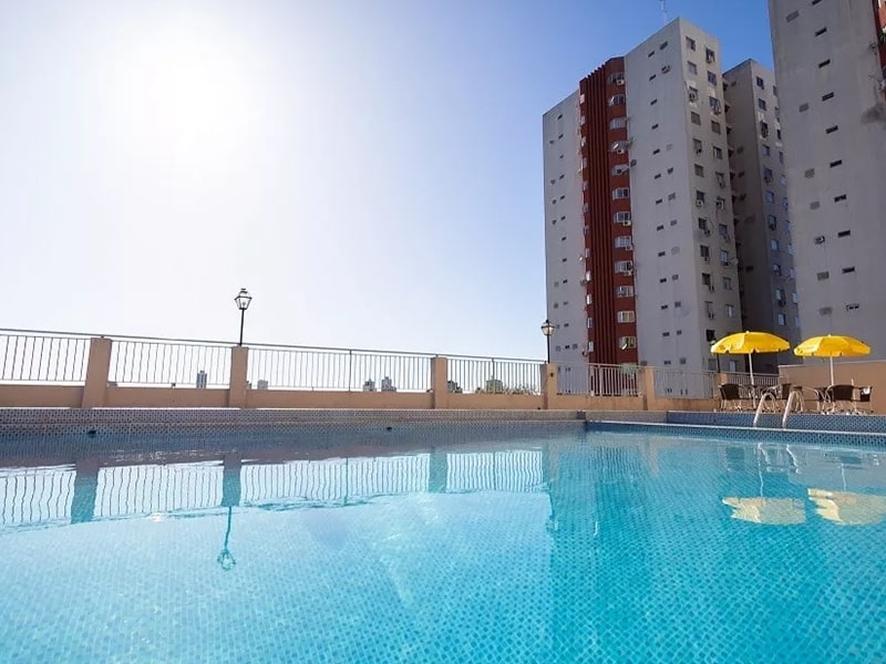 Piscina azul ao ar livre com cadeiras e guarda-sóis, cercada por prédios.