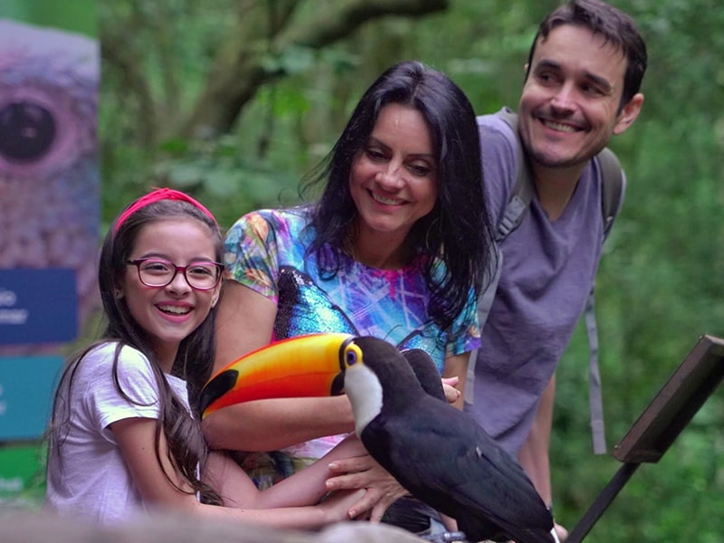 Família sorridente interagindo com um tucano em uma experiência de contato com a natureza no Parque das Aves, representando a diversão e o encantamento das atrações em Foz do Iguaçu.